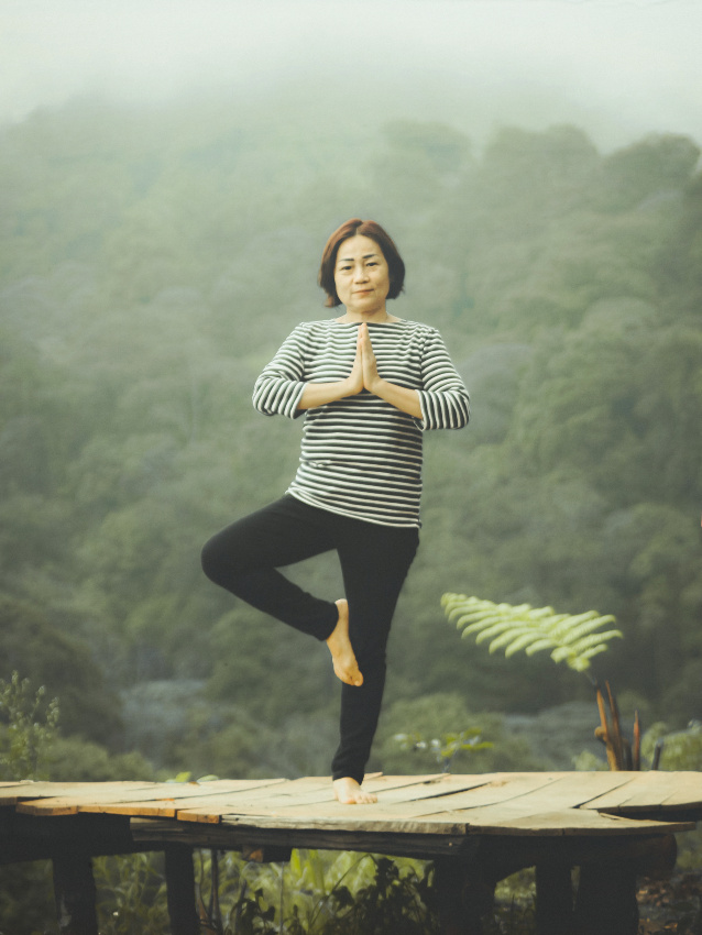 Asiatische Frau in der Baum-Pose des Yoga auf einer Holzbrücke mit Urwald im Hintergrund.